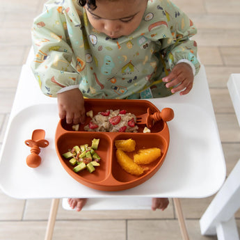 Toddler uses Bumkins Silicone Grip Dish: Clay, enjoying finger foods on a divided orange plate with suction on the high chair tray.