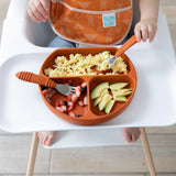 Baby in a high chair, wearing an orange bib, enjoys eggs and fruit from the Bumkins Silicone Grip Dish: Clay with suction base.