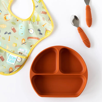 Yellow bib with camping print, Bumkins Silicone Grip Dish: Clay, and two orange-handled utensils on a white background.