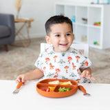 A child beams at the table with assorted foods on a Bumkins Silicone Grip Dish: Clay, paired with a colorful bib.