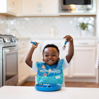 A smiling child with a Bumkins Spoon + Fork: Dark Blue set enjoys their meal in the kitchen, confidently using the silicone utensils.