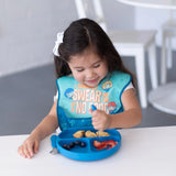 A young girl in a bib happily eats with the Bumkins Spoon + Fork: Dark Blue set in a bright room.