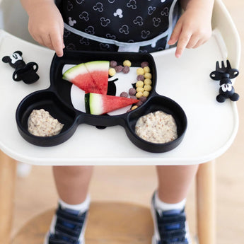 Toddler feeding is easy with Bumkins Mickey Mouse Silicone Grip Dish holding watermelon, cereal balls, and porridge on the high chair.