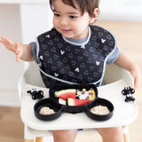 A toddler uses Bumkins Mickey Mouse Silicone Chewtensils® and a Mickey-shaped plate for baby-led weaning, wearing a Mickey-patterned bib.