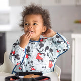 Child with curly hair wearing a Bumkins Sleeved Bib: Mickey Mouse, munching snacks in a high chair.