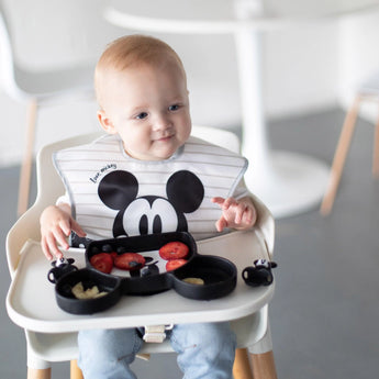 A toddler in a highchair enjoys strawberries and bananas from a Bumkins Mickey Mouse Silicone Grip Dish with a matching bib.