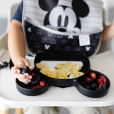 A toddler in a high chair enjoys bowtie pasta and berries using a Mickey Mouse utensil, with a Bumkins Silicone Grip Dish: Mickey Mouse and bib.