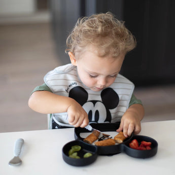 A toddler joyfully eats strawberries, grapes, and breaded food from a Bumkins Silicone Grip Dish: Mickey Mouse using a fork.