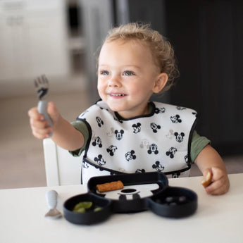 A curly-haired toddler with a Mickey Mouse Silicone Grip Dish by Bumkins uses a fork at the table, wearing a matching bib.