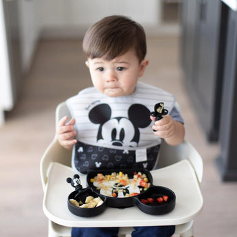 Toddler in a high chair, wearing a Mickey Mouse bib, holds a toy and eats from Bumkins Silicone Grip Dish: Mickey Mouse.