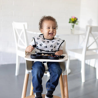 Toddler in a high chair, smiling with food from a Bumkins Silicone Grip Dish: Mickey Mouse, wearing jeans and a patterned bib.