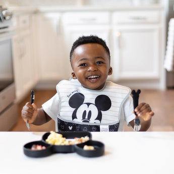 A toddler in a high chair eagerly holds utensils over a Bumkins Silicone Grip Dish: Mickey Mouse, kitchen in the background.