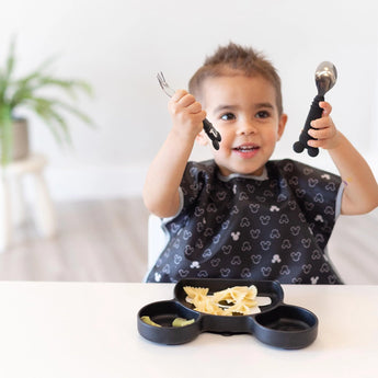 A smiling child eagerly grips a fork and spoon, ready to enjoy pasta from their Bumkins Silicone Grip Dish: Mickey Mouse, with a plant behind.