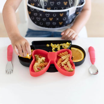 A child enjoys colorful pasta from a Bumkins Minnie Mouse Silicone Grip Dish, with corn and oranges, using red utensils and wearing a bib.