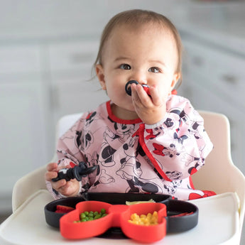 A toddler wears a patterned bib and uses a hand-shaped fork to eat from Bumkins Minnie Mouse Silicone Grip Dish with suction base.