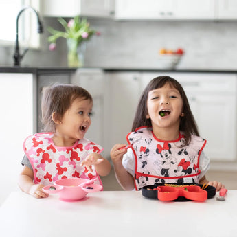 Two kids in Minnie Mouse bibs enjoy their meal with Bumkins Silicone Grip Dish: Minnie Mouse to keep food secure at the table.