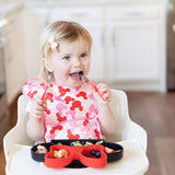 A toddler in a high chair eats using Bumkins Minnie Mouse fork, spoon, and Silicone Grip Dish with suction base while wearing a pink bib.