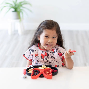 Young girl in a Minnie Mouse-themed outfit smiles by a table with Bumkins Silicone Grip Dish: Minnie Mouse and fork, snacks in front.