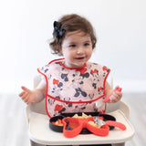 A toddler in a bow sits in a high chair with a Bumkins Minnie Mouse bib, eating from a black and red Silicone Grip Dish.