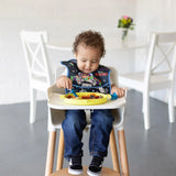 A toddler self-feeds from a Bumkins Silicone Grip Dish: Toy Story Alien, in a high chair with a space-themed bib, white chairs nearby.