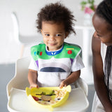 Curly-haired child in high chair, wearing a space bib, holds Bumkins Silicone Grip Dish: Toy Story Alien. Smiling woman next to them.