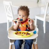 A toddler with curly hair sits in a high chair, wearing a colorful bib. They hold a blue utensil and have a Bumkins Silicone Grip Dish featuring Toy Story Alien filled with pasta and berries. A white chair and table are in the background.