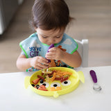 A toddler self-feeds from a Bumkins Toy Story Alien dish, focused on using a purple fork with colorful pasta and fruit.