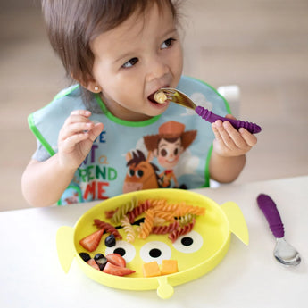 A toddler self-feeds pasta and fruit from a Toy Story Alien silicone dish, wearing a bib with cartoon characters.