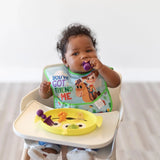 Baby in a high chair eagerly self-feeds from a Bumkins Silicone Grip Dish: Toy Story Alien while wearing a bib and using a spoon.