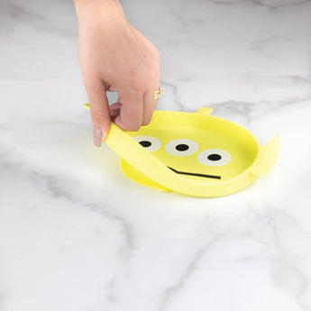 A hand peels a Bumkins Silicone Grip Dish: Toy Story Alien with eyes and a mouth, perfect for toddler self-feed adventures.