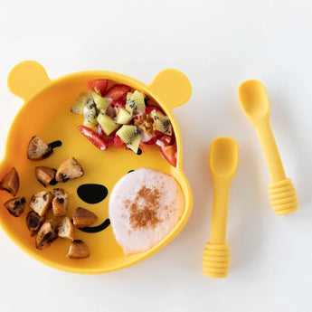 The Bumkins Silicone Grip Dish: Winnie The Pooh holds fruit, toast, and yogurt with two yellow baby spoons on a white surface.