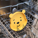 The Bumkins Silicone Grip Dish: Winnie The Pooh, a food-safe yellow bear plate with a smile, is in the dishwasher rack.