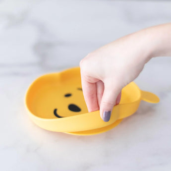 A hand holds a Bumkins Winnie The Pooh Silicone Grip Dish in yellow on a white surface.