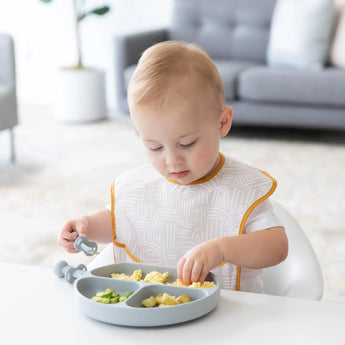 A toddler enjoys a meal from a Bumkins Silicone Grip Dish in Gray at a table in a bright, modern living room.