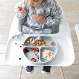 A baby in a high chair enjoys food from a Bumkins Silicone Grip Dish: Gray, paired with a zigzag bib and secure suction base.