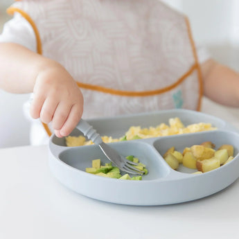 A child enjoys veggies with a fork from a Bumkins Silicone Grip Dish: Gray, featuring a suction base, while wearing a bib.