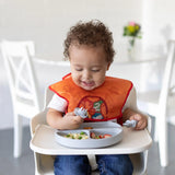 A curly-haired toddler in an orange bib smiles in a high chair, enjoying food from a Bumkins Silicone Grip Dish: Gray with a fork.