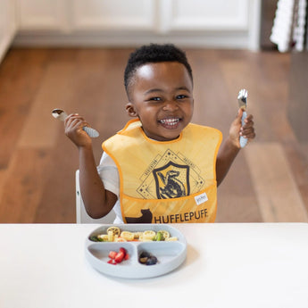 A child in a Hufflepuff bib and Bumkins Gray Silicone Grip Dish smiles, using a fork and spoon with colorful food in front.