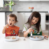 Two young kids in high chairs eat with forks on Bumkins Gray Silicone Grip Dishes. Bright bibs add color to the sunny kitchen.