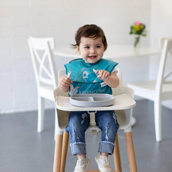 Toddler in a high chair, blue bib on, joyfully holding a spoon with a Bumkins Silicone Grip Dish: Gray on the white table behind.