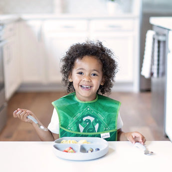 A curly-haired child in a green bib beams at the table, enjoying a meal from a gray Bumkins Silicone Grip Dish in the bright kitchen.