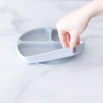 Hand lifting Bumkins Silicone Grip Dish: Gray with toddler-friendly suction base from a marble surface.