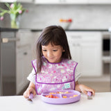 A young girl in a colorful bib sits at the table with a purple plate, excited to use her Bumkins Spoon + Fork: Lavender set.