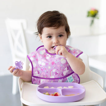 A baby with a purple bib enjoys their meal from a Bumkins Lavender Silicone Grip Dish, featuring a secure suction base.