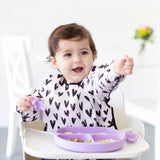 A smiling toddler in a high chair, wearing a heart-patterned bib, reaches for food from Bumkins Silicone Grip Dish: Lavender.