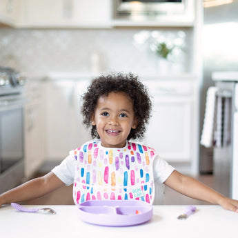 A toddler smiles in a bright kitchen wearing a colorful bib with Bumkins Lavender Silicone Grip Dish and utensils with suction base.