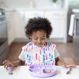 A child, wearing a colorful bib, smiles at their meal with Bumkins Lavender Spoon + Fork while a purple plate sits nearby.