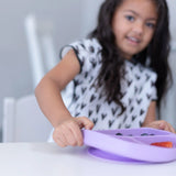 A child reaching for a Bumkins Silicone Grip Dish in Lavender, filled with blueberries and a strawberry slice on a white table.