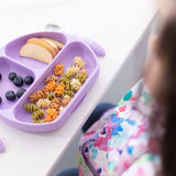 A child enjoys colorful pasta, apple slices, and blueberries on a Bumkins Silicone Grip Dish: Lavender, in a charming floral outfit.