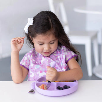 Young girl with long hair enjoys her meal from a Bumkins Lavender Silicone Grip Dish, wearing a purple unicorn shirt and a white bow.